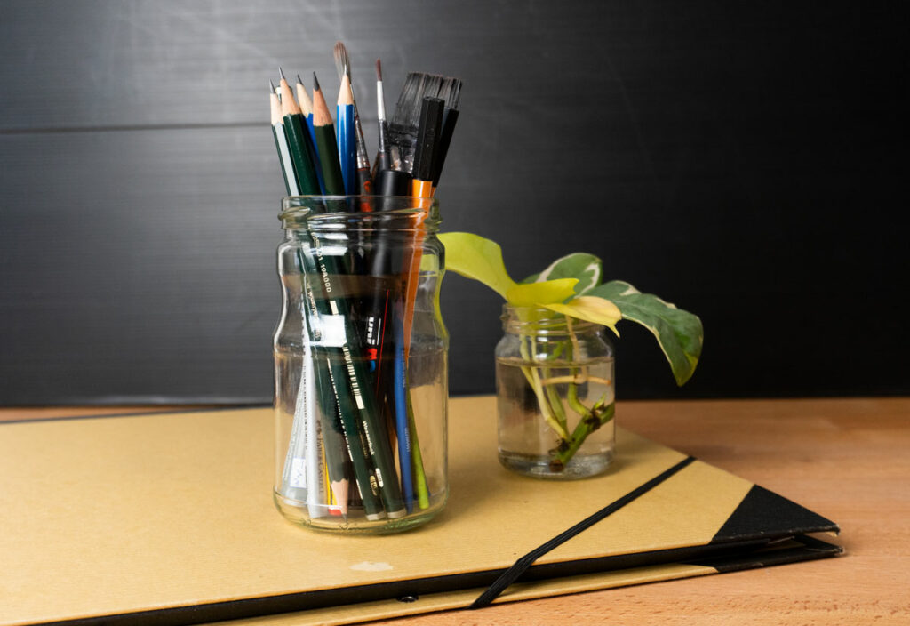 Eine Mappe auf der ein Glas mit Stiften und Pinseln steht, sowie eine Wasserglas mit Efeututen-Ablegern.