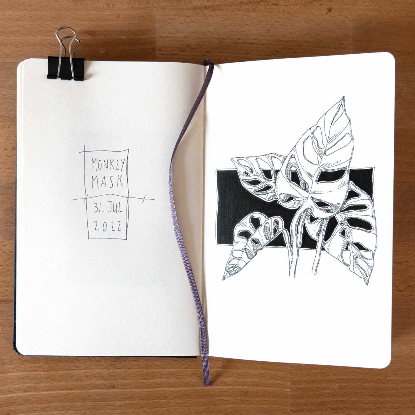 Geöffnetes Skizzenbuch mit gewebtem Bändchen und Papierklemme. Auf der Linkenseite der Schriftzug "Monkey Mask 31. Juli 2022" und auf der rechten die Zeichnung von meiner Monkey Mask.
